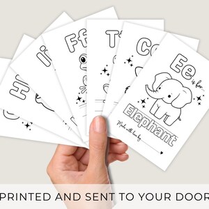 May include: A hand holding a stack of white flashcards with black line drawings and text. Each card features a letter of the alphabet and an associated image, such as an elephant. The text "PRINTED AND SENT TO YOUR DOOR" is at the bottom.