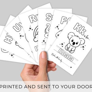 Puede incluir: Una mano sostiene un abanico de tarjetas blancas con dibujos lineales negros de animales y letras. La tarjeta superior dice "KK is for... Koala" con una ilustraci&oacute;n de koala. El texto "Made with love by" es visible. La parte inferior de la imagen dice "PRINTED AND SENT TO YOUR DOOR".