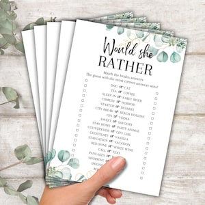 May include: A stack of white paper game cards with the text "Would she RATHER" in black and gold. The cards have a floral design with eucalyptus leaves. The game includes questions like "Dog or Cat" and "Tea or Coffee".