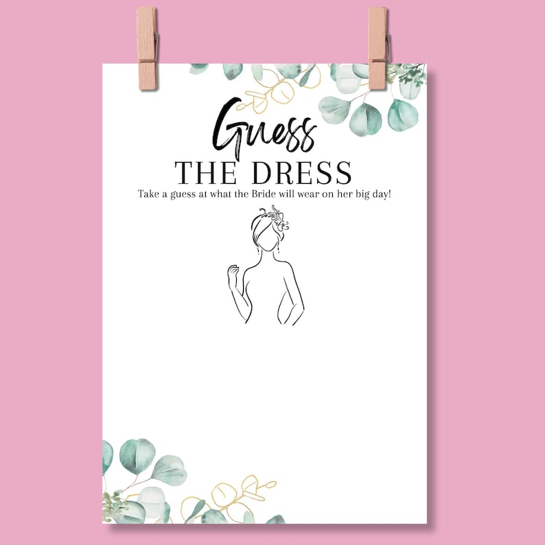May include: A white paper sign with the text "Guess the Dress" in black script. The sign includes a line drawing of a bride and is decorated with watercolor eucalyptus leaves and gold accents. The sign is held by two wooden clothespins.