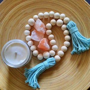 May include: A wooden bead garland with two turquoise tassels and three pink Himalayan salt rocks on a wooden plate.