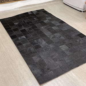 Puede incluir: Una alfombra negra rectangular con un diseño de patchwork. La alfombra está hecha de piezas cuadradas de material, creando una superficie texturizada. Está colocada sobre un suelo de madera clara.