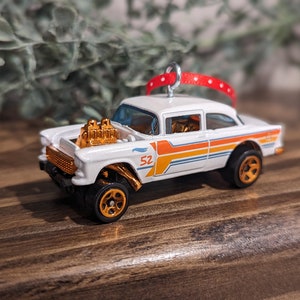 May include: White 1955 Chevrolet with orange and white racing stripes, gold engine, and gold wheels. The car is a Christmas ornament with a red ribbon.