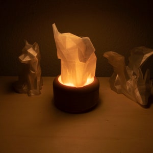 Op de afbeelding: Drie witte geometrische dierenbeelden, een vos, een olifant en een draak, verlicht door een warm licht van een kaars in de olifantensculptuur.