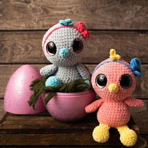 May include: Two crocheted bird toys, one gray and one pink, with large googly eyes. The gray bird is sitting in a pink egg-shaped container. The pink bird is sitting on a wooden crate.