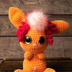 May include: Orange crocheted bunny toy with pink and white fur on its head, large black eyes, and pink accents on its ears.