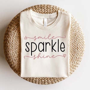 以下が含まれることがあります： 白いTシャツに、「smile sparkle shine」という文字が黒とピンクの文字で書かれています。Tシャツは折り畳まれて、籐製の敷物の上に置かれています。