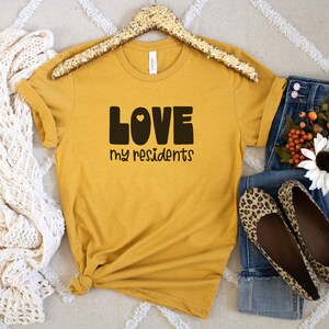 K&ouml;nnte beinhalten: Ein senfgelbes T-Shirt mit dem Text "LOVE my residents" in schwarzen Blockbuchstaben mit einem kleinen Herzen im "O".