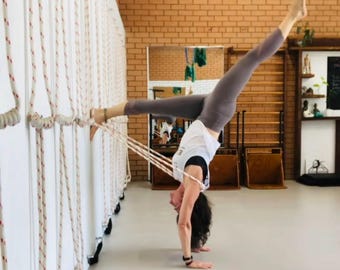 May include: A person performing a handstand with legs split in a yoga studio. The person is wearing a white top and gray leggings, using ropes attached to a wall for support. The studio has a brick wall and a white wall with ropes.