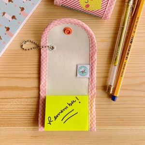 May include: A collection of stationery items including a pink and white gingham notepad, a small lemon-themed pouch, and a memo holder with a yellow sticky note that says "Remember!". A gold pen and a blue pen are also present.