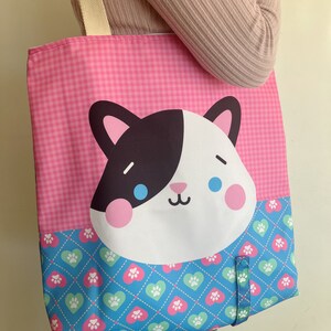 May include: Pink and blue tote bag with a cartoon cat face on the front. The bag has a checkered pattern on the top half and a pattern of hearts and paw prints on the bottom half.