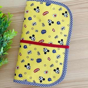 Könnte beinhalten: Gelbe Stoff-Federmappe mit Mickey-Maus-Druck. Die Federmappe hat ein rotes Gummiband und einen blau-weiß karierten Besatz.