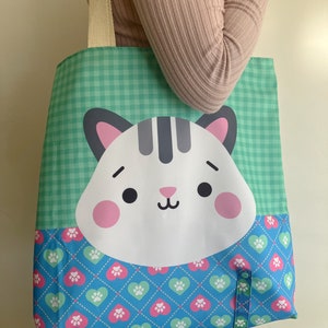 May include: A green and blue tote bag featuring a white cat face and paw print design. The bag has a cream-coloured handle.