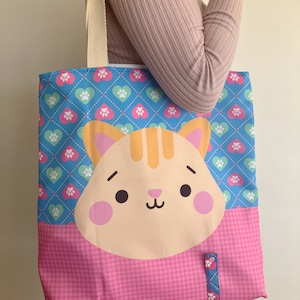 May include: A blue and pink tote bag with a cute cartoon cat face on the front. The bag has a white handle and a blue and white patterned strap.