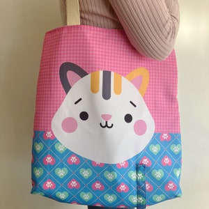 May include: Pink tote bag featuring a cartoon cat face. The upper section has a pink gingham pattern, and the lower section has a blue diamond pattern with heart and paw print designs. It has a beige strap.