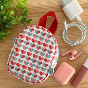Puede incluir: Una pequeña bolsa redonda con cremallera, de color rojo y azul con estampado de corazones, con un asa roja. La bolsa está sobre una superficie de madera con un cargador blanco, un par de auriculares blancos, una funda rosa para auriculares y un pintalabios rosa.