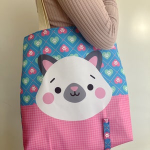 May include: A tote bag with a cartoon cat face, pink cheeks, and a grey nose. The top section has a blue diamond pattern with pink and green paw print hearts. The bottom is pink gingham. It has cream-coloured straps.