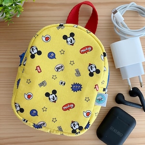 May include: A small yellow backpack with a Mickey Mouse print. The backpack has a red handle and a blue zipper. The backpack is on a wooden surface with a white phone charger, a white phone cable, and a black case for wireless earbuds.