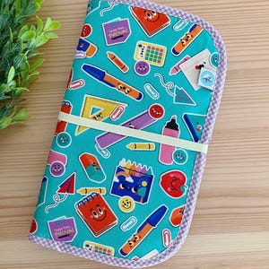 Könnte beinhalten: Ein türkisfarbener Stiftetui mit einem violetten und weißen Karomuster. Das Etui ist mit einem Muster aus Schulutensilien bedeckt, darunter Bleistifte, Buntstifte, Radiergummis und ein Lineal. Der Text "Tergi-ken PROVA!" wird auf dem Etui wiederholt.