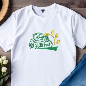 Pode incluir: Camiseta branca com um trator verde e um desenho de trigo amarelo.