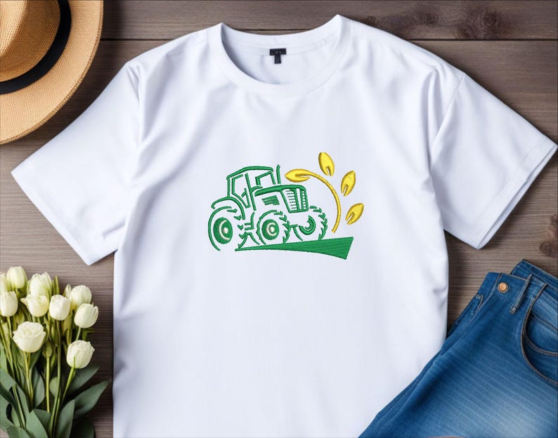 Pode incluir: Camiseta branca com um trator verde e um desenho de trigo amarelo.