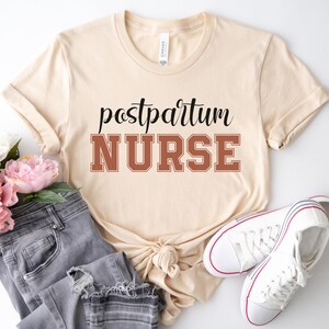 May include: A beige t-shirt with the text "postpartum NURSE" in black and brown lettering. The shirt is lying on a white surface with a pair of white trainers and a pair of blue jeans.