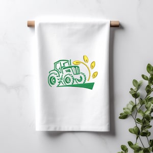 Pode incluir: Uma toalha de cozinha branca com um desenho bordado verde de um trator dirigindo por um campo de trigo amarelo.