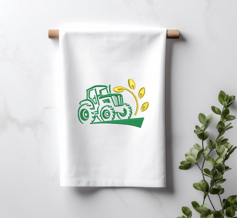 Pode incluir: Uma toalha de cozinha branca com um desenho bordado verde de um trator dirigindo por um campo de trigo amarelo.
