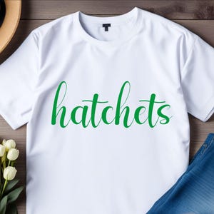 May include: White t-shirt with the word "hatchets" written in green cursive lettering.
