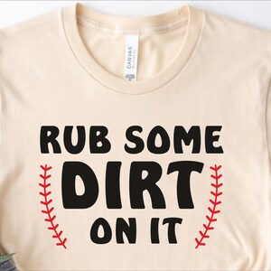 May include: A beige t-shirt with the text "RUB SOME DIRT ON IT" in black lettering. The text is surrounded by two red baseball stitch designs.