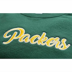 Puede incluir: Una sudadera de cuello redondo en color verde oscuro con la palabra Packers bordada en una tipografía cursiva amarilla con borde blanco en el pecho. La tela tiene una apariencia suave y texturizada, con costuras claramente visibles.