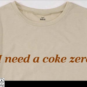 May include: A beige t-shirt with the text "I need a coke zero" printed in brown on the front.