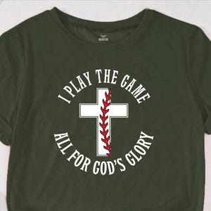 May include: A green t-shirt with a white circle design featuring a baseball with a red stitch and a white cross. The text inside the circle reads "I Play The Game All For God's Glory".