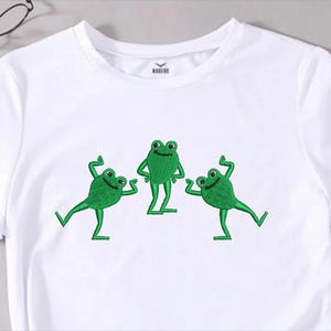 May include: White t-shirt featuring an embroidered design of three green frogs. The frogs have smiling faces and are in various poses. The shirt has short sleeves and a crew neck. A pair of eyeglasses is visible in the background.