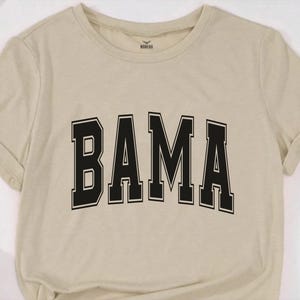 May include: A beige t-shirt with the word "BAMA" printed in black block letters.