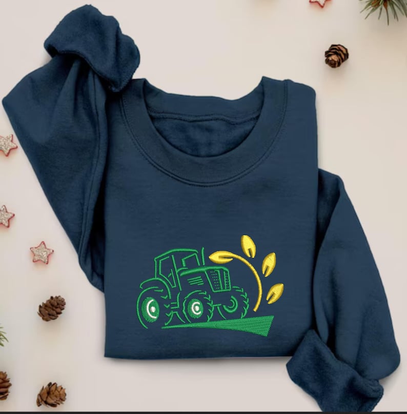 Pode incluir: Uma camisola azul marinho com um design bordado em verde e amarelo de um trator a puxar um caule de trigo.