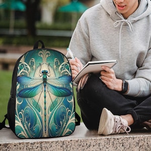 Pode incluir: Uma mochila preta com um gráfico grande e detalhado de uma libélula azul e verde centrado no painel frontal. A libélula está rodeada por intrincados padrões de filigrana cor de creme sobre um fundo azul profundo.