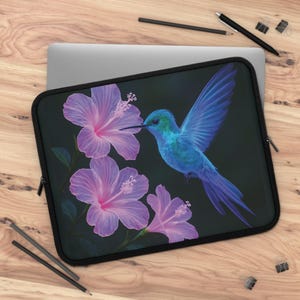 May include: Black laptop sleeve featuring a vibrant blue hummingbird with outstretched wings, hovering near three pink hibiscus flowers. The design is set against a dark background, creating a striking contrast. The sleeve has a black zipper and is designed to protect a laptop.