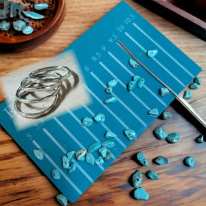 May include: Four silver rings stacked on a turquoise blue ring sizing chart with numbers 5, 5.5, 6, 6.5, 7, 7.5, 8, 8.5, 9, 9.5, 10, and 10.5. Turquoise stones are scattered around the chart.