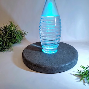Könnte beinhalten: Eine klare Glasflasche mit wellenförmigem Design, beleuchtet mit blauem Licht, steht auf einem dunkelgrauen, runden Steinsockel. Grüne Zweige mit Blättern sind auf beiden Seiten angeordnet und schaffen eine minimalistische, moderne Ästhetik.