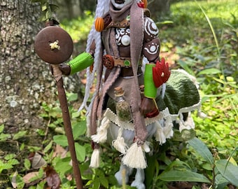 May include: A detailed doll figure with dark skin, white hair, and elaborate tribal-inspired clothing. The doll holds a staff with a wooden button and a glowing orange orb. The figure is adorned with green, brown, and white accents, and stands in a natural outdoor setting.