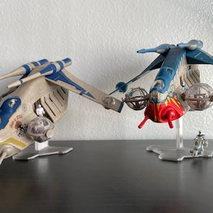 Peut inclure: Deux vaisseaux spatiaux Star Wars en jouet sur des supports d'exposition. Le vaisseau spatial de gauche est blanc, bleu et jaune avec les mots "Obi-Wan's Star" imprimés sur le côté. Le vaisseau spatial de droite est bleu, rouge et blanc.