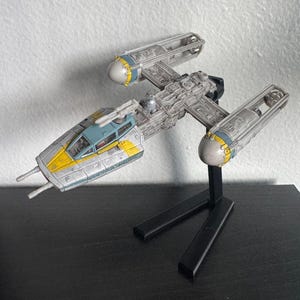 Könnte beinhalten: Detailliertes Modell eines Y-Wing-Sternjägers mit grau-gelbem Farbschema. Das Modell ist auf einem schwarzen Ständer montiert, der sein kompliziertes Design und seine Details zeigt. Das Cockpit ist blau und gelb.