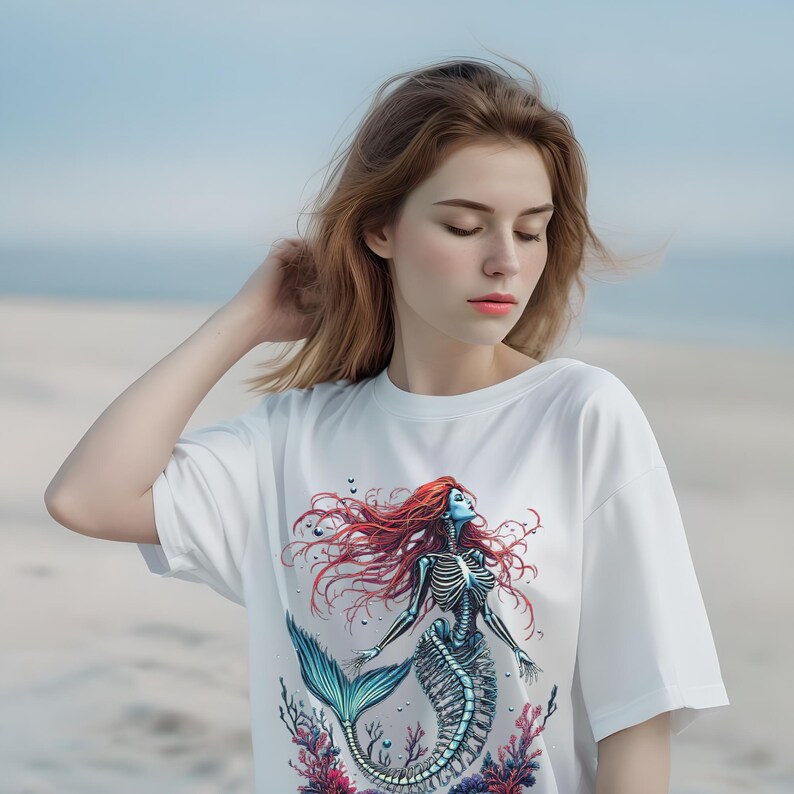 May include: White t-shirt featuring a skeletal mermaid design with red hair, a blue face, and a teal tail. The graphic includes coral and seaweed elements. The shirt is a casual, short-sleeved style.