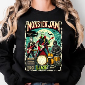 May include: Black sweatshirt featuring a Monster Jam concert poster design. The graphic depicts a band of monsters playing instruments, with Frankenstein on guitar. The text reads "Monster Jam", "The Crypt Club", and "Live One Night".