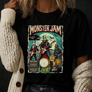 May include: Black t-shirt featuring a band of monsters playing instruments under a full moon. The band includes Frankenstein on guitar, a vampire on guitar, and a gorilla on drums. The text "Monster Jam" and "Live" are also visible.