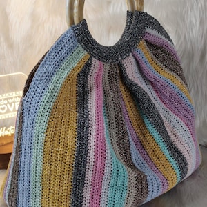 May include: A handmade crochet tote bag with a striped pattern in various colors, including blue, yellow, pink, and silver. The bag features a rounded shape and two marbled, light-colored handles. The bag's design is a mix of textures and colors.