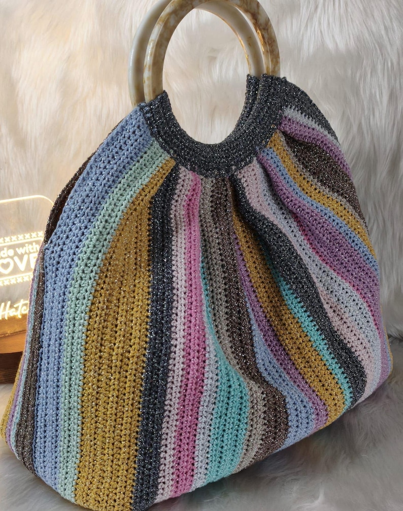 May include: A handmade crochet tote bag with a striped pattern in various colors, including blue, yellow, pink, and silver. The bag features a rounded shape and two marbled, light-colored handles. The bag's design is a mix of textures and colors.
