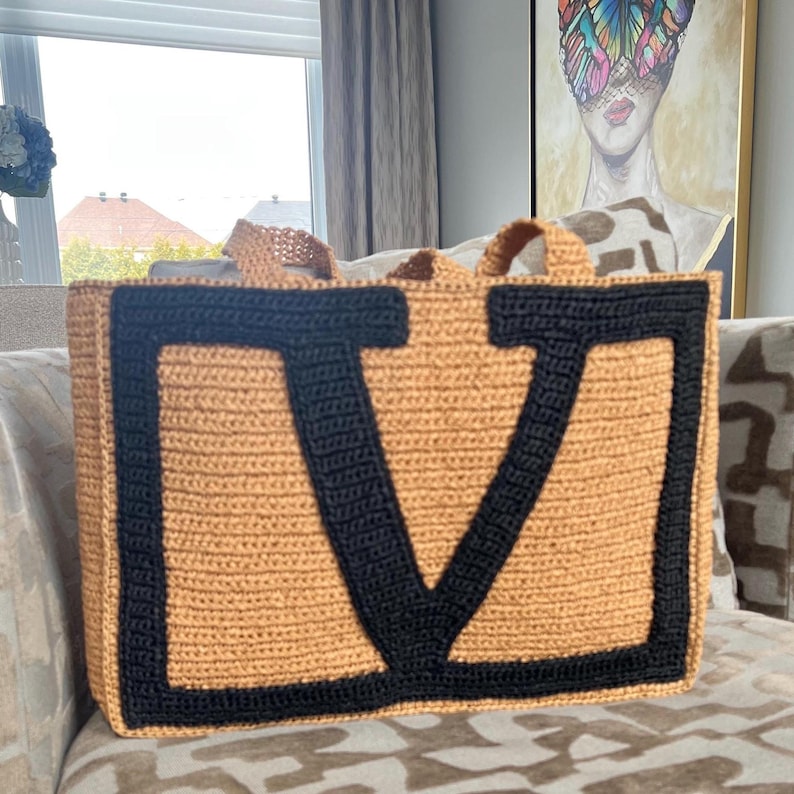 May include: A rectangular crochet tote bag featuring a tan base color with a prominent, large black letter V stitched onto the front. The bag includes two matching tan crochet handles at the top, showcasing a textured, hand-crafted knit construction.