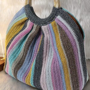 May include: A handmade crochet handbag with a striped pattern in various colors including yellow, pink, blue, and gray. The bag has a round, marbled handle and a structured shape, perfect for carrying essentials. The bag is sitting on a white, fluffy surface.